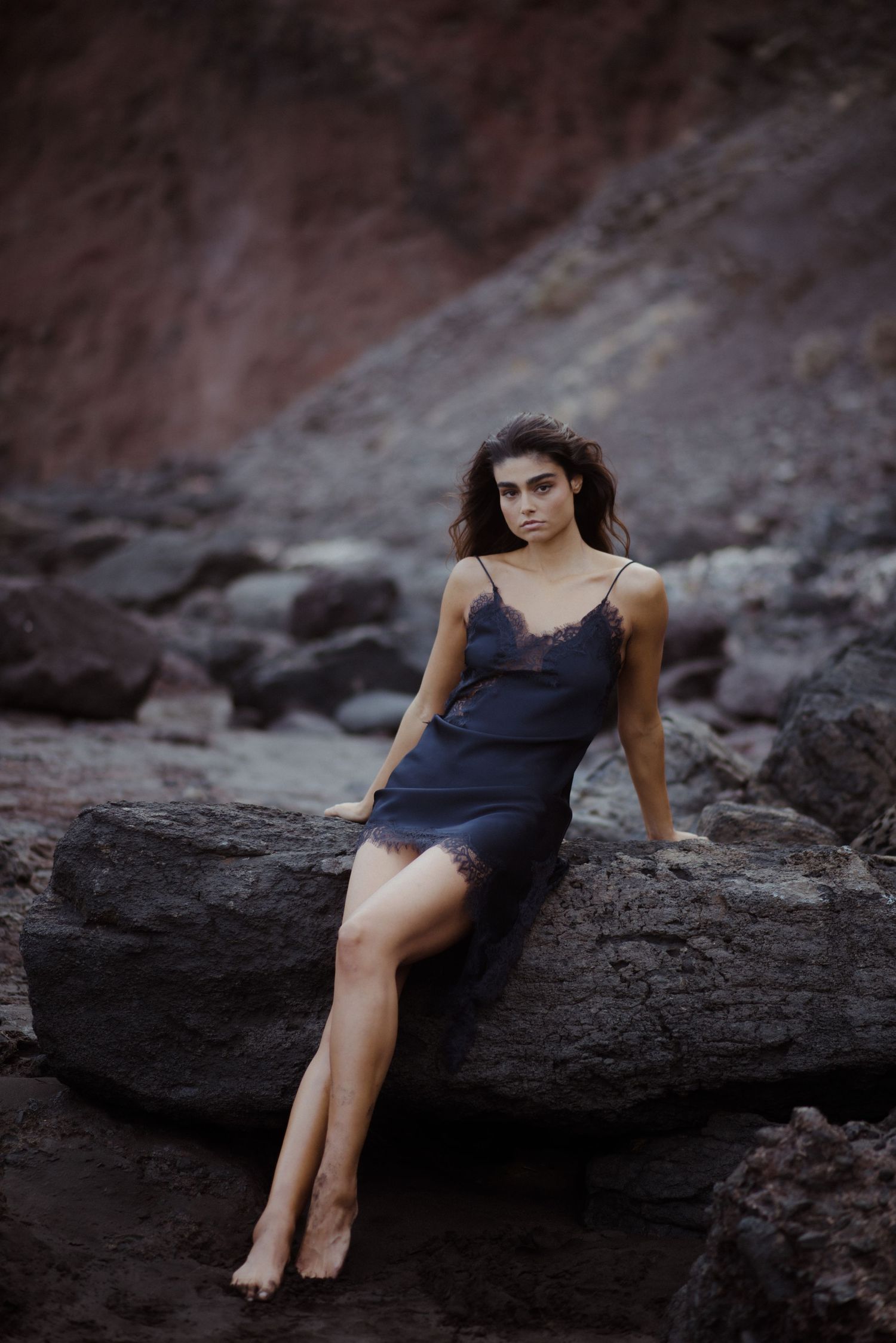 A woman in a navy blue dress poses gracefully on dark rocky terrain at sunset.
