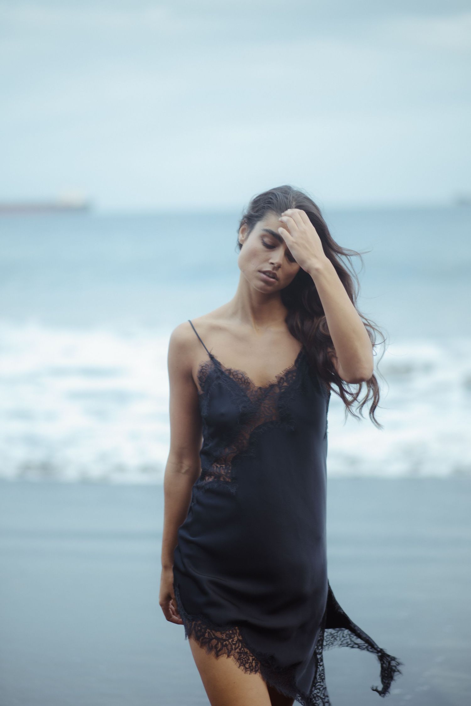 A figure in a black lace camisole poses expressively against a misty ocean backdrop in a moody photoshoot.