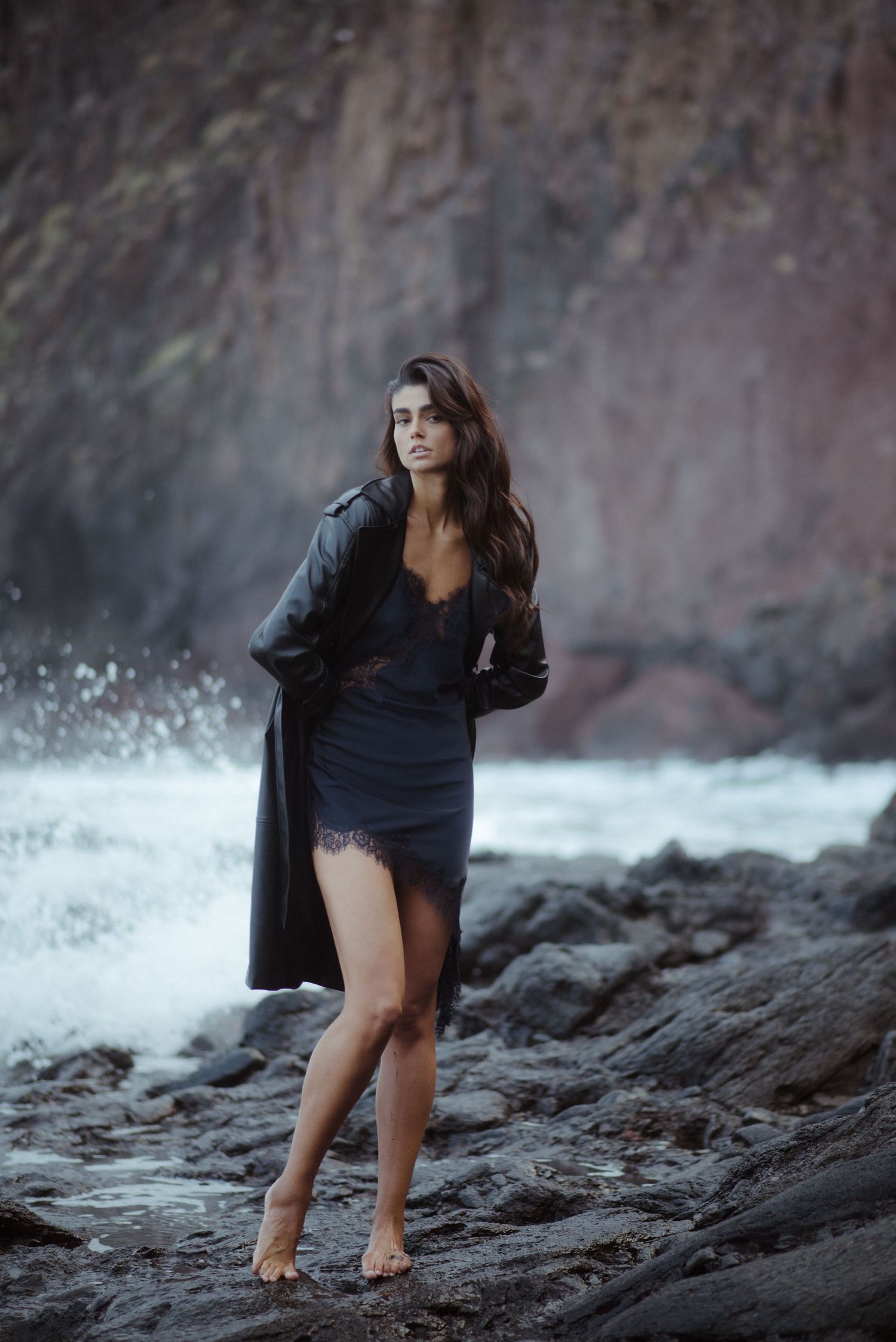Woman in dark dress poses gracefully on rocky coastal shoreline with crashing waves.