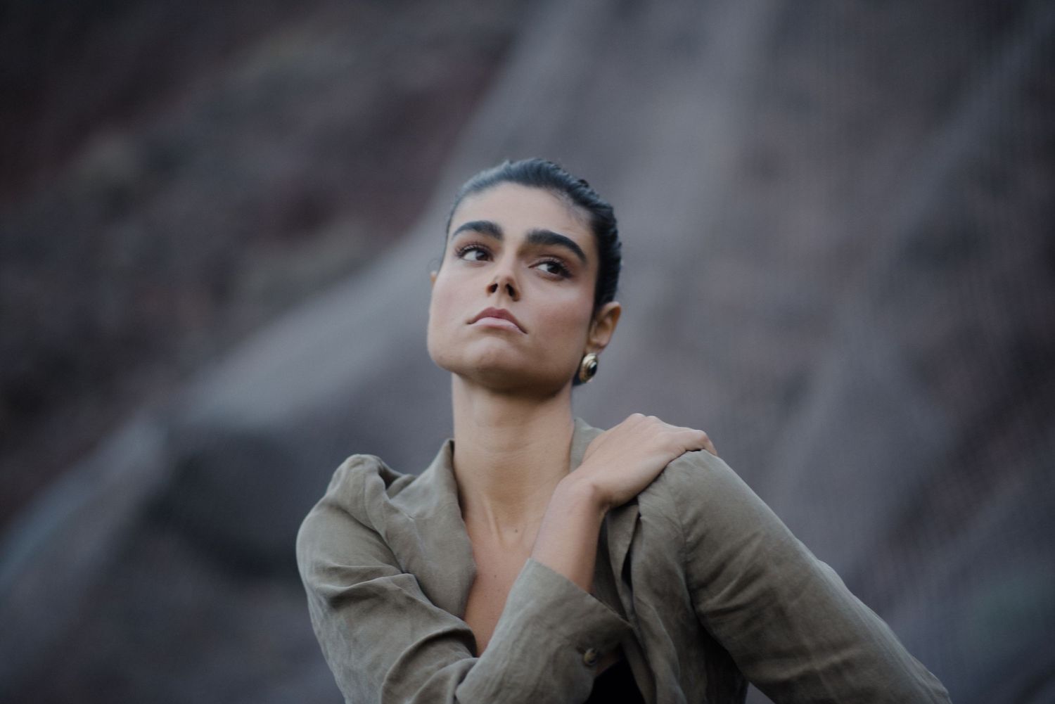 Person in olive colored outfit poses against dark rocky background in dramatic lighting.