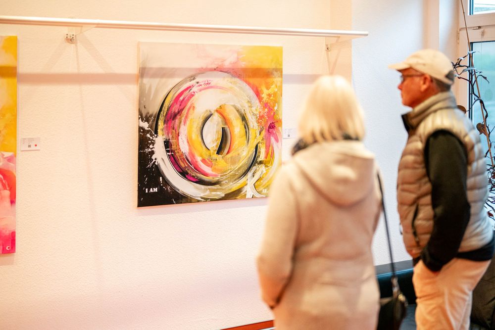 Gallery visitors observe a vibrant abstract circular artwork with yellow and black swirls displayed on a beige wall.