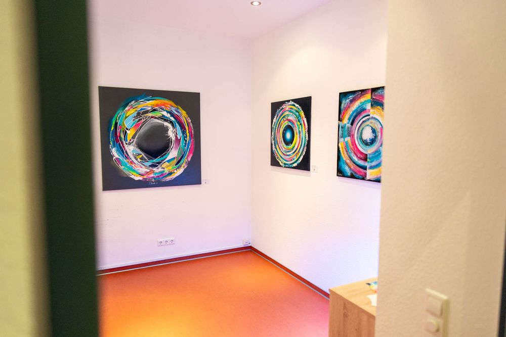 Abstract circular artwork with vibrant colors displayed on white walls in a gallery-like setting.