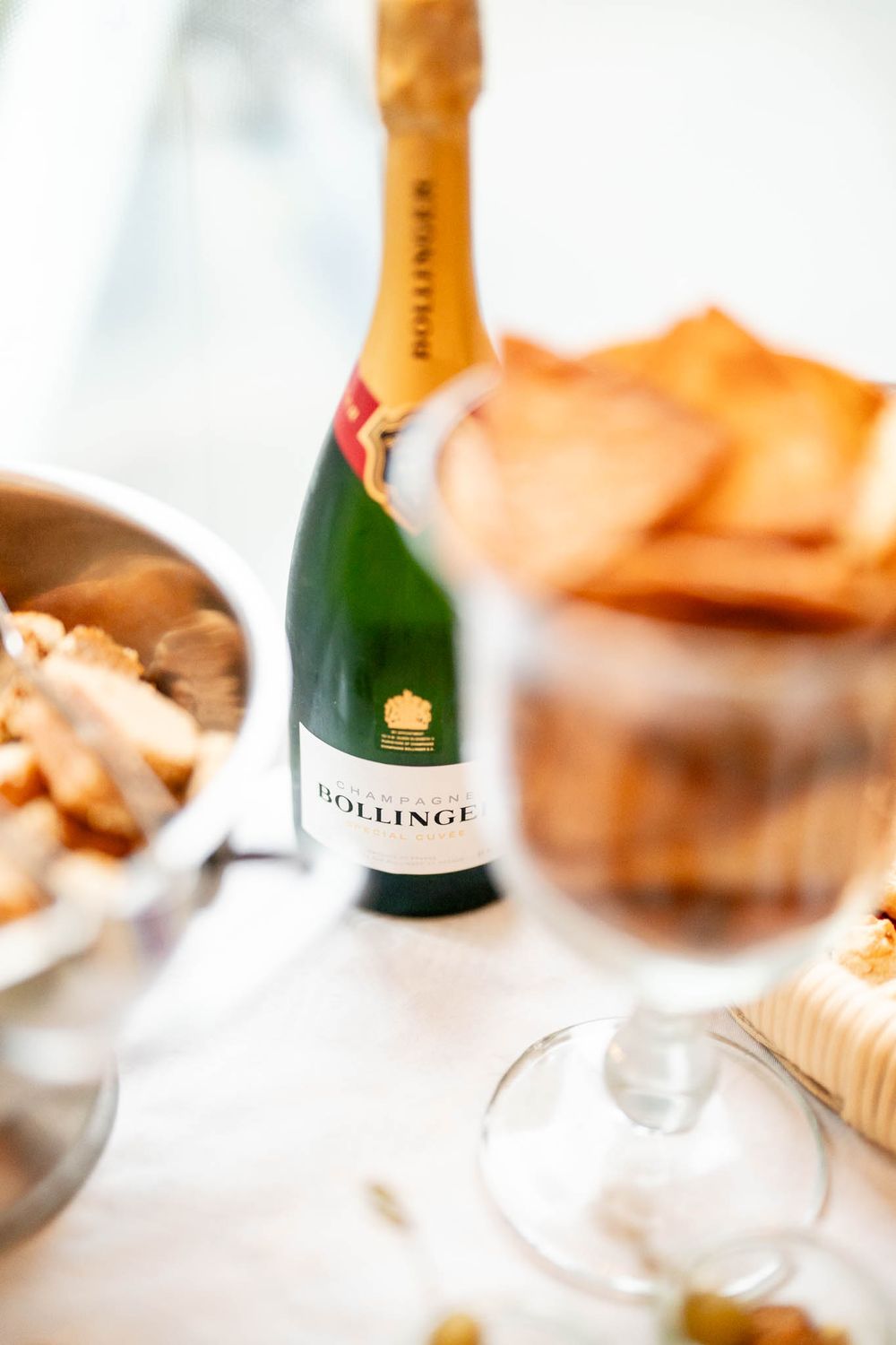 A bottle of Bollinger champagne sits on a white table with wine glasses and snacks in soft focus.