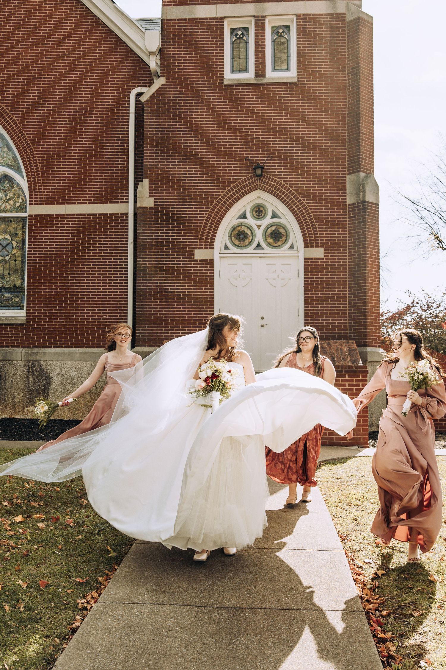 Bride twirling in white gown with bridesmaids in pink dresses outside a red brick church.