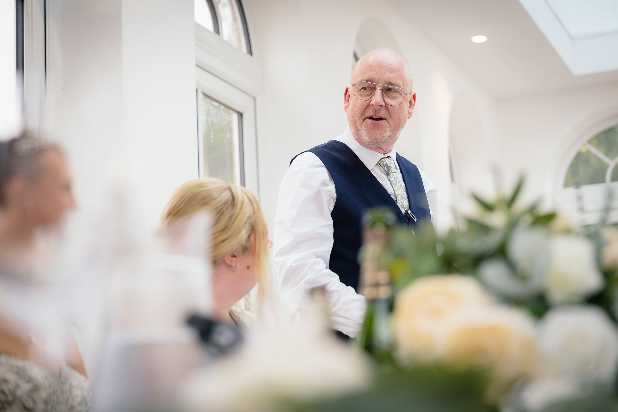 Wedding guests enjoying a celebration in a bright conservatory-style venue with natural lighting. Woodhill Hall.