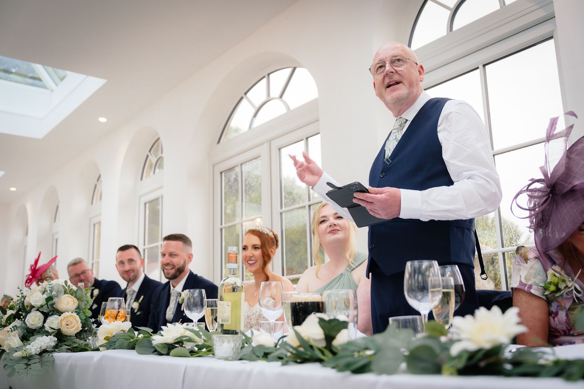 Wedding reception venue decorated with floral arrangements and natural light streaming through arched windows. Woodhill Hall.