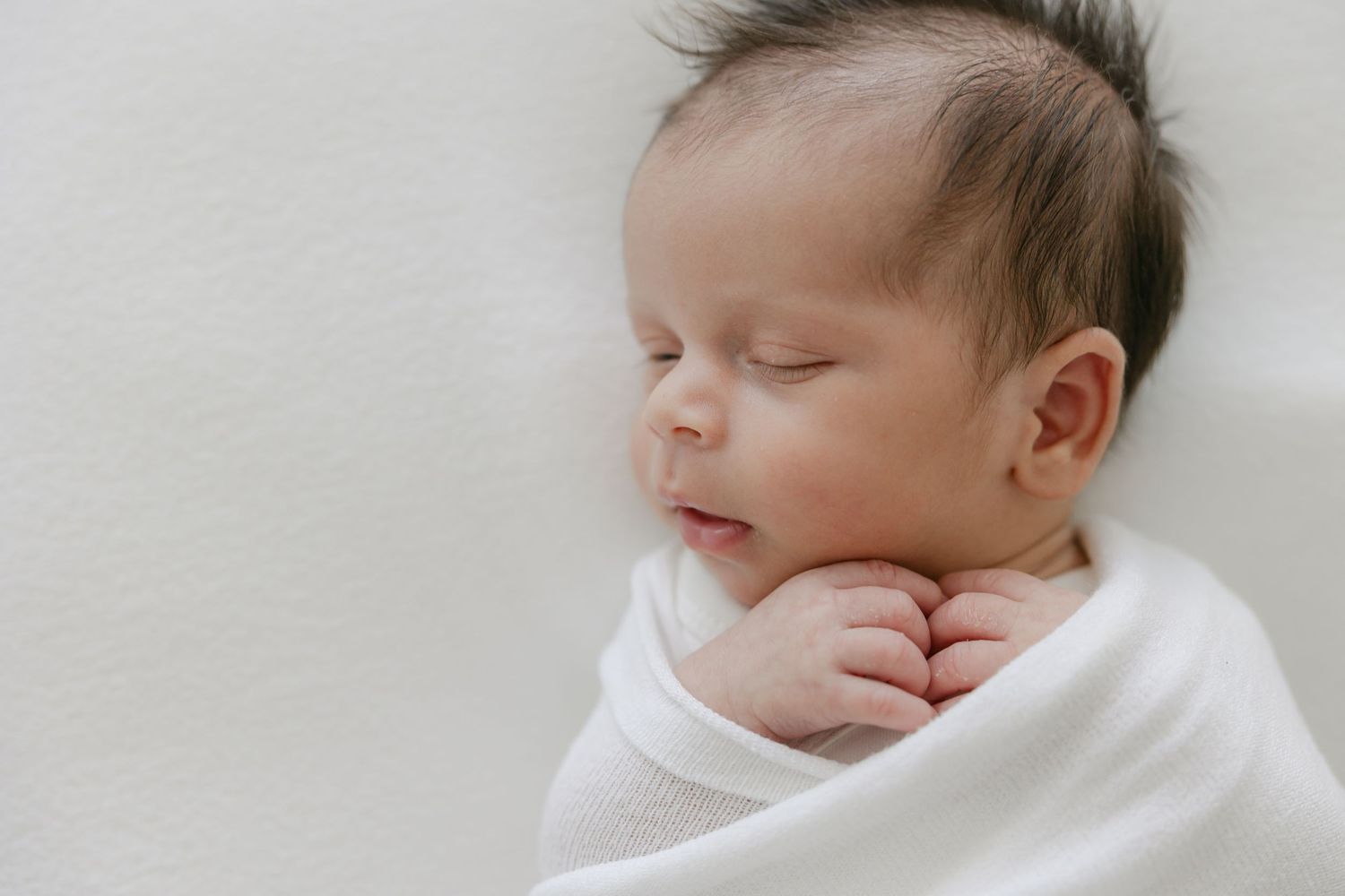 A sleeping newborn baby wrapped in white swaddle blanket rests peacefully against light background.