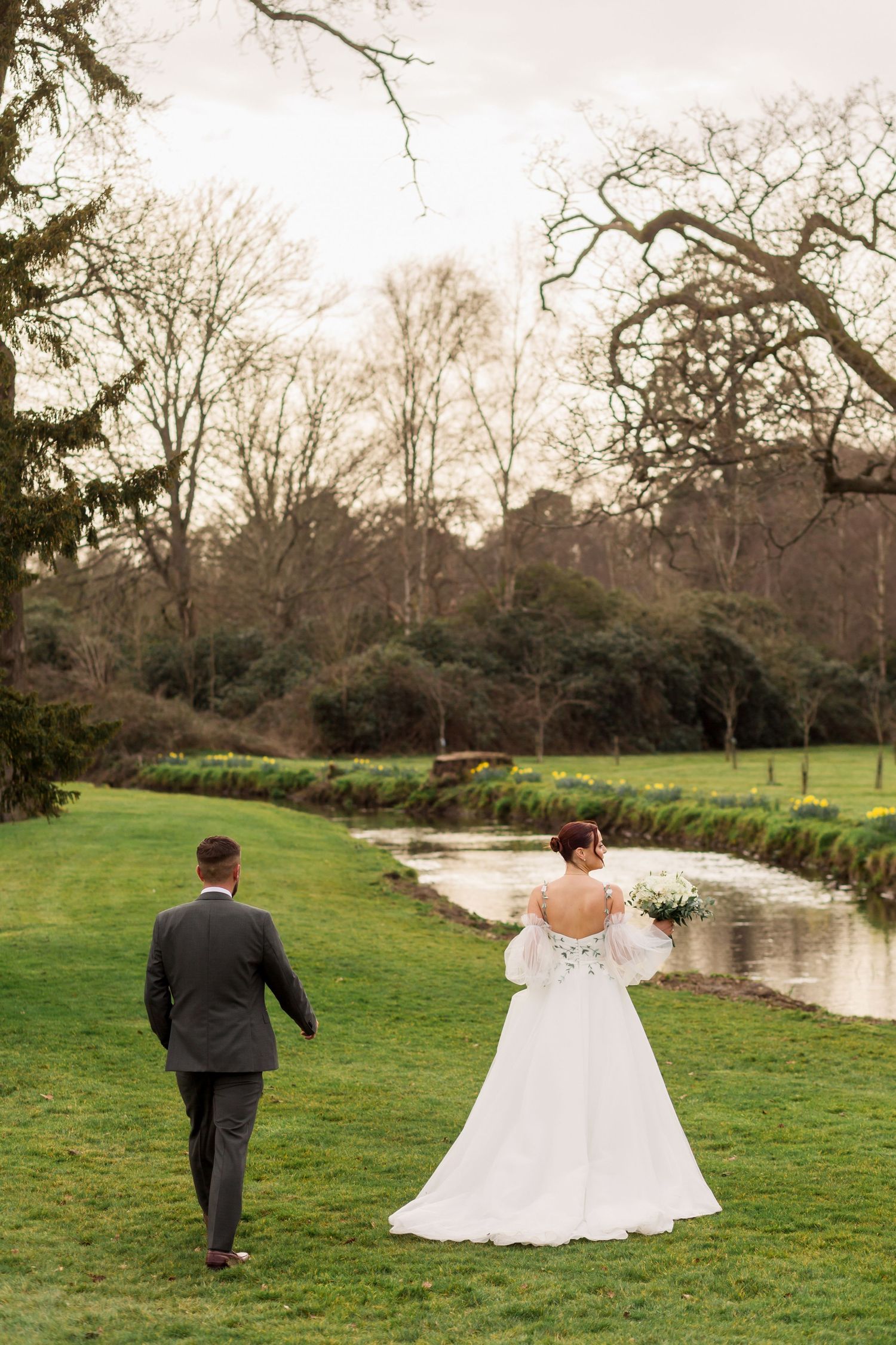 A couple walks hand in hand across a lush green lawn beside a winding stream on their wedding day.