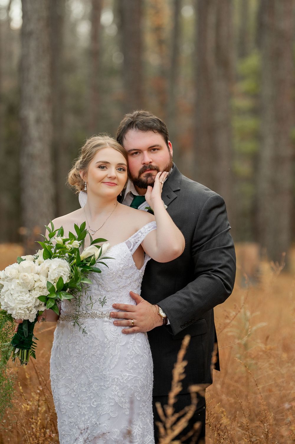 Wedding couple poses in rustic woodland setting with white floral bouquet.