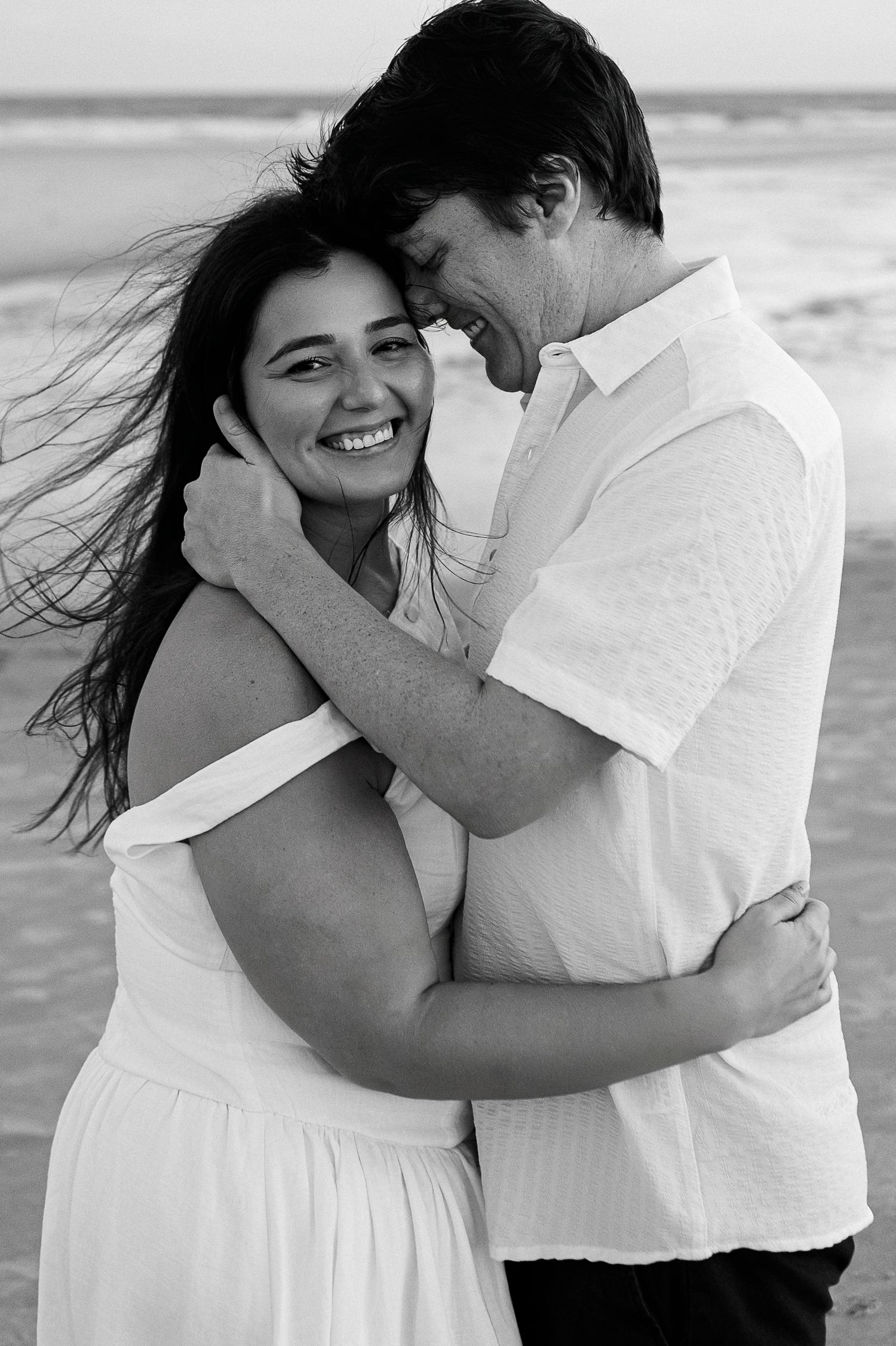 Black and white beach portrait of laughing couple in white clothing embracing by the ocean.