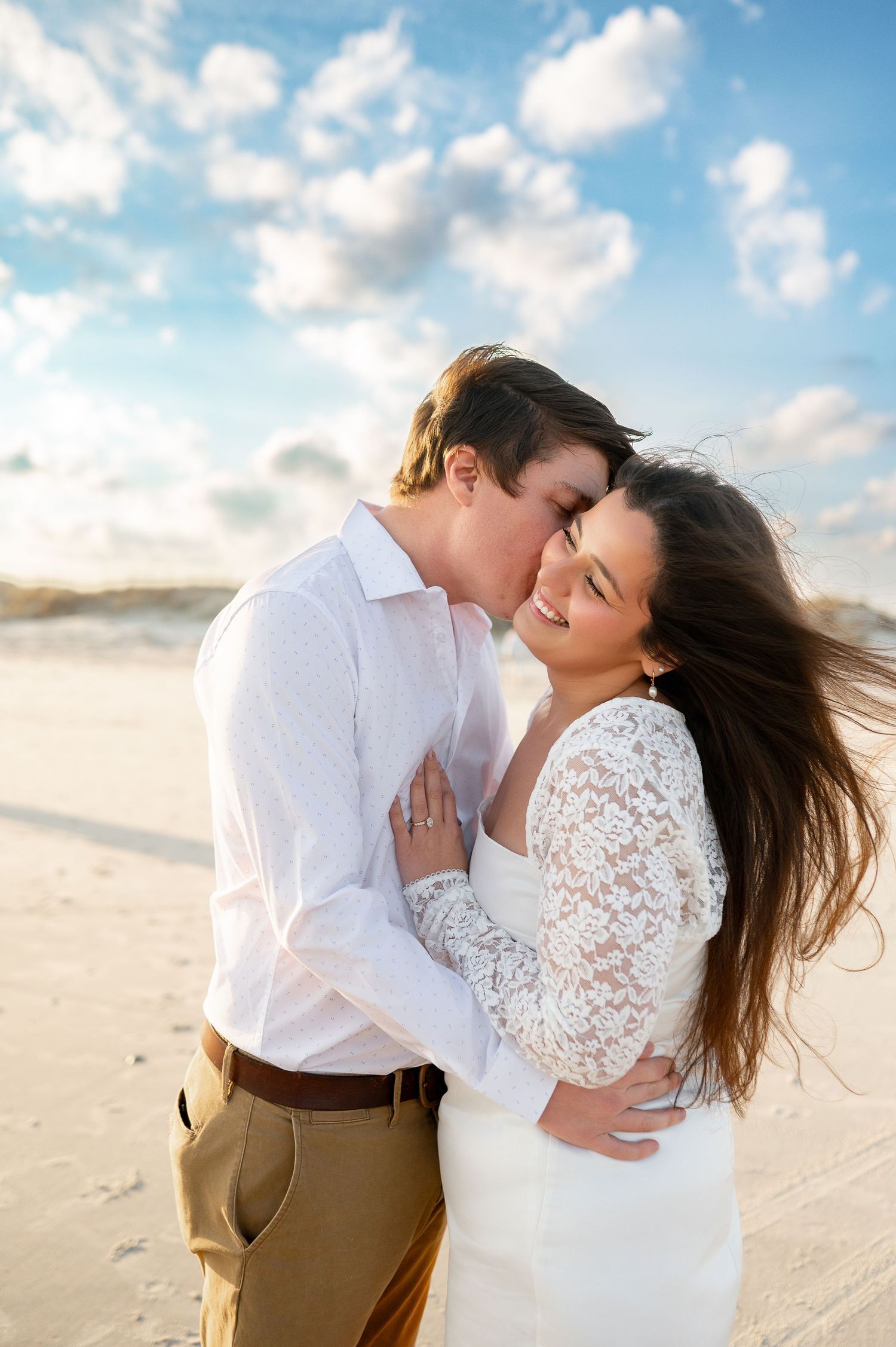 Couple sharing intimate moments on sandy beach with blue sky and clouds in background.