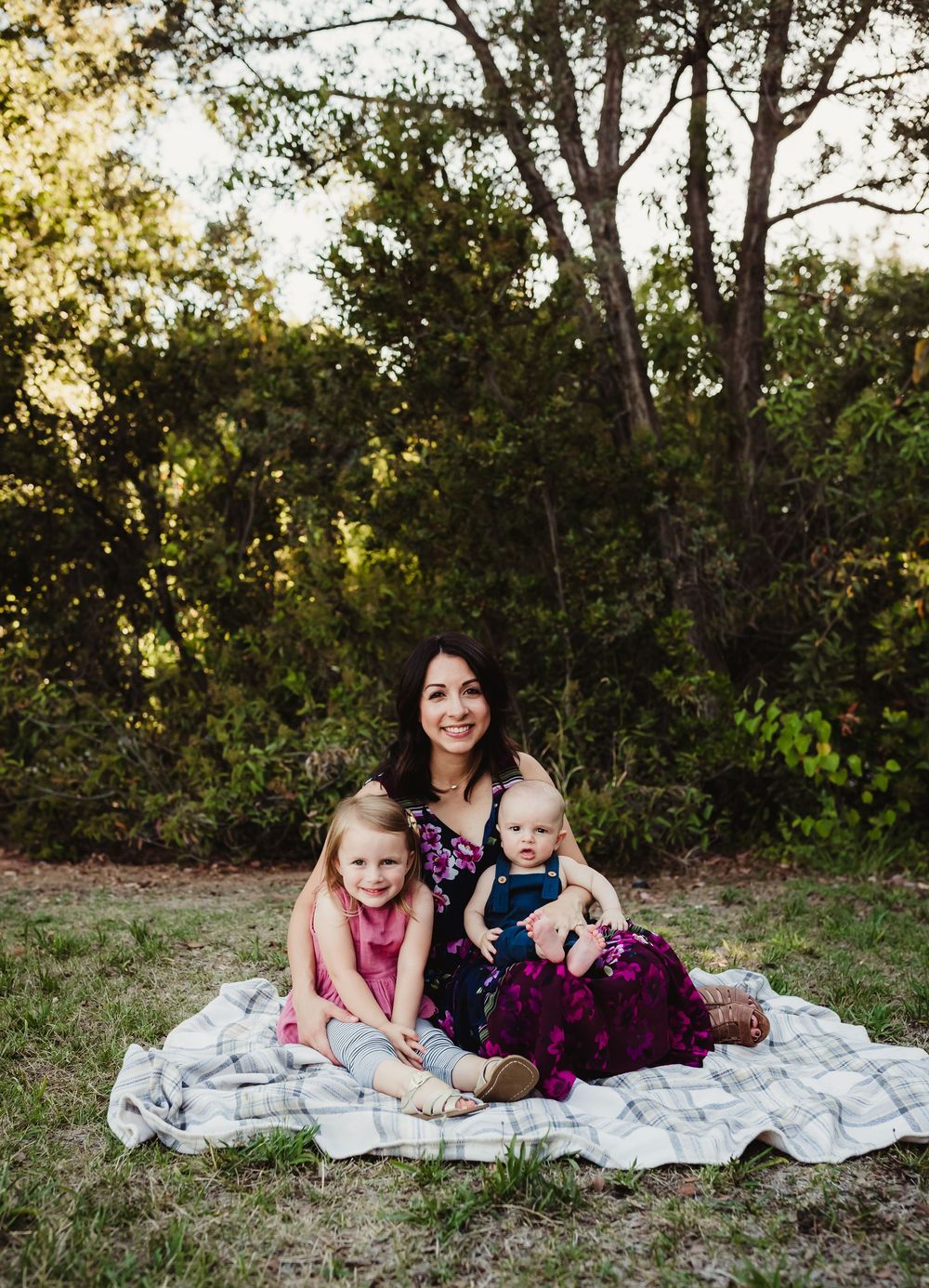 A family sits together on a blanket outdoors during golden hour with trees and greenery in the background.