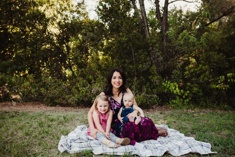 A group relaxes together on a blanket in a sunlit wooded area during an outdoor photo session.