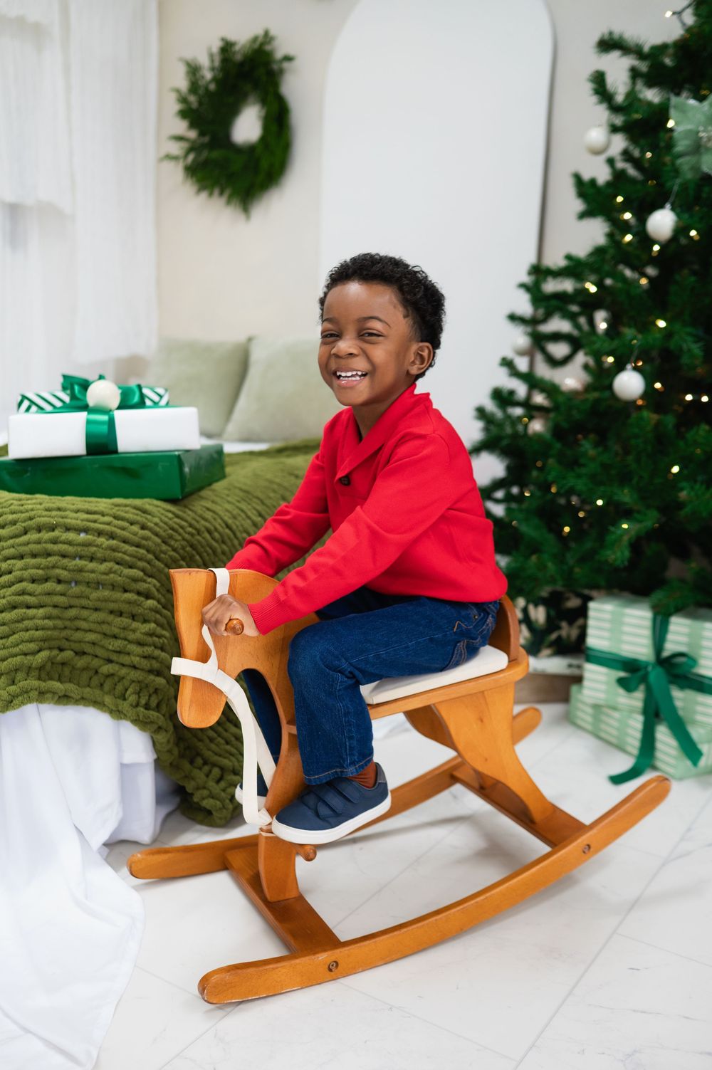 smiling kid on rocking horse at lume and sage photo studio during christmas mini session; tree, wreath, and presents in background.