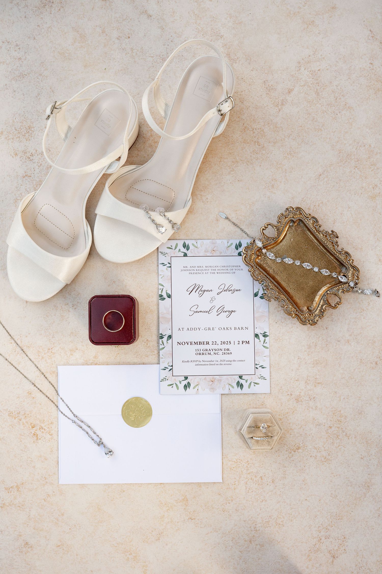 Wedding details including white shoes, invitations, jewelry and ring box arranged on a beige textured surface.