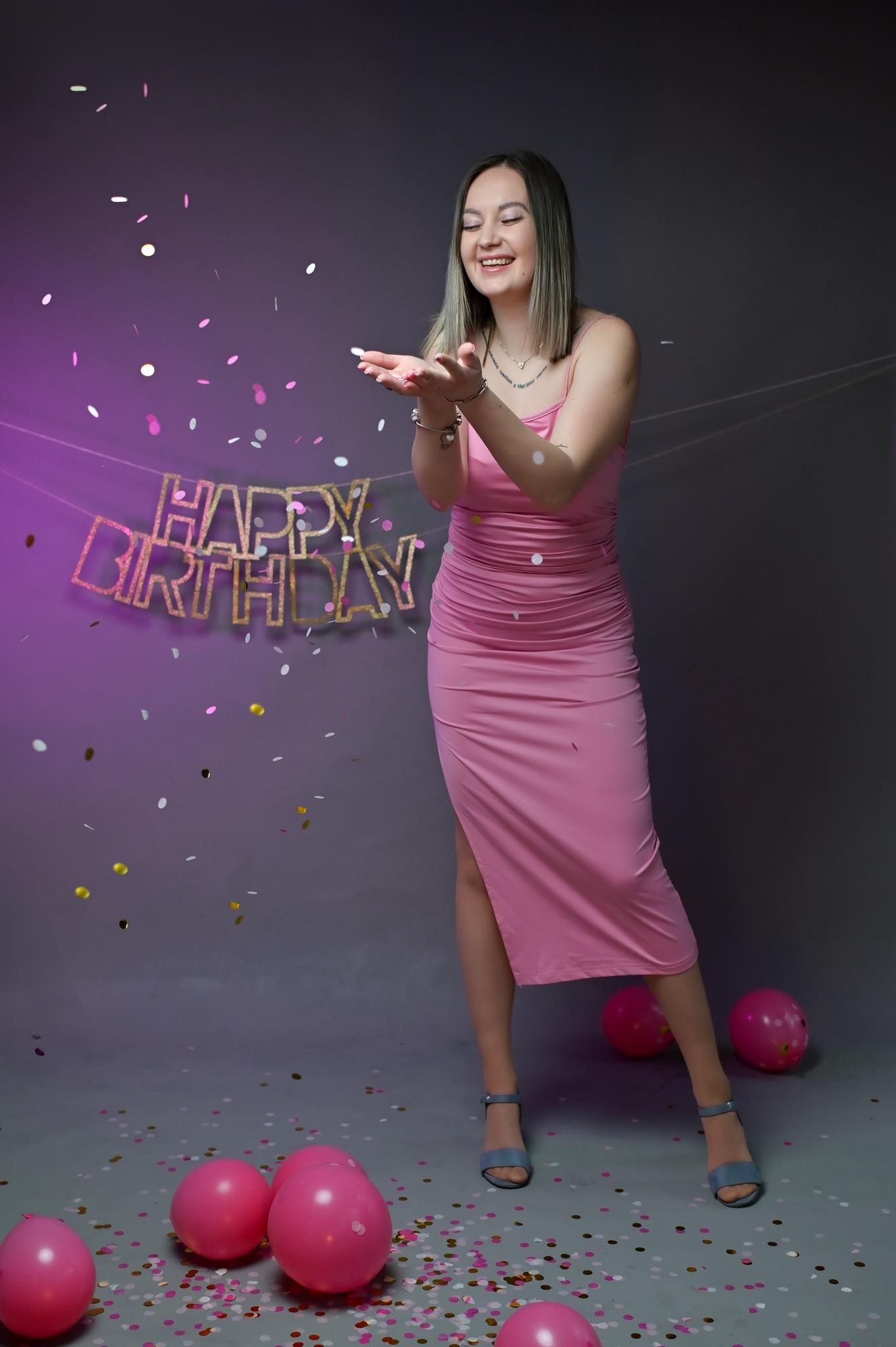 Person in pink dress celebrates with confetti and pink balloons against dark backdrop during birthday party photoshoot.