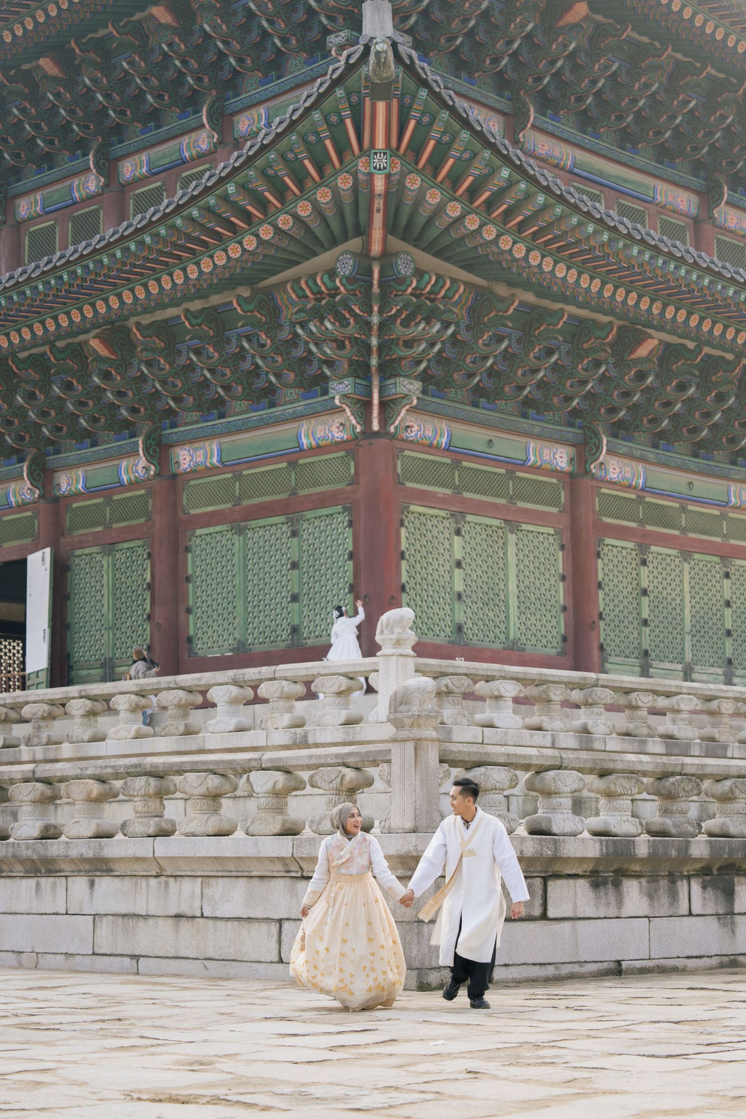 Traditional Korean hanok building with ornate roof details and two people sitting on stone steps in traditional dress.