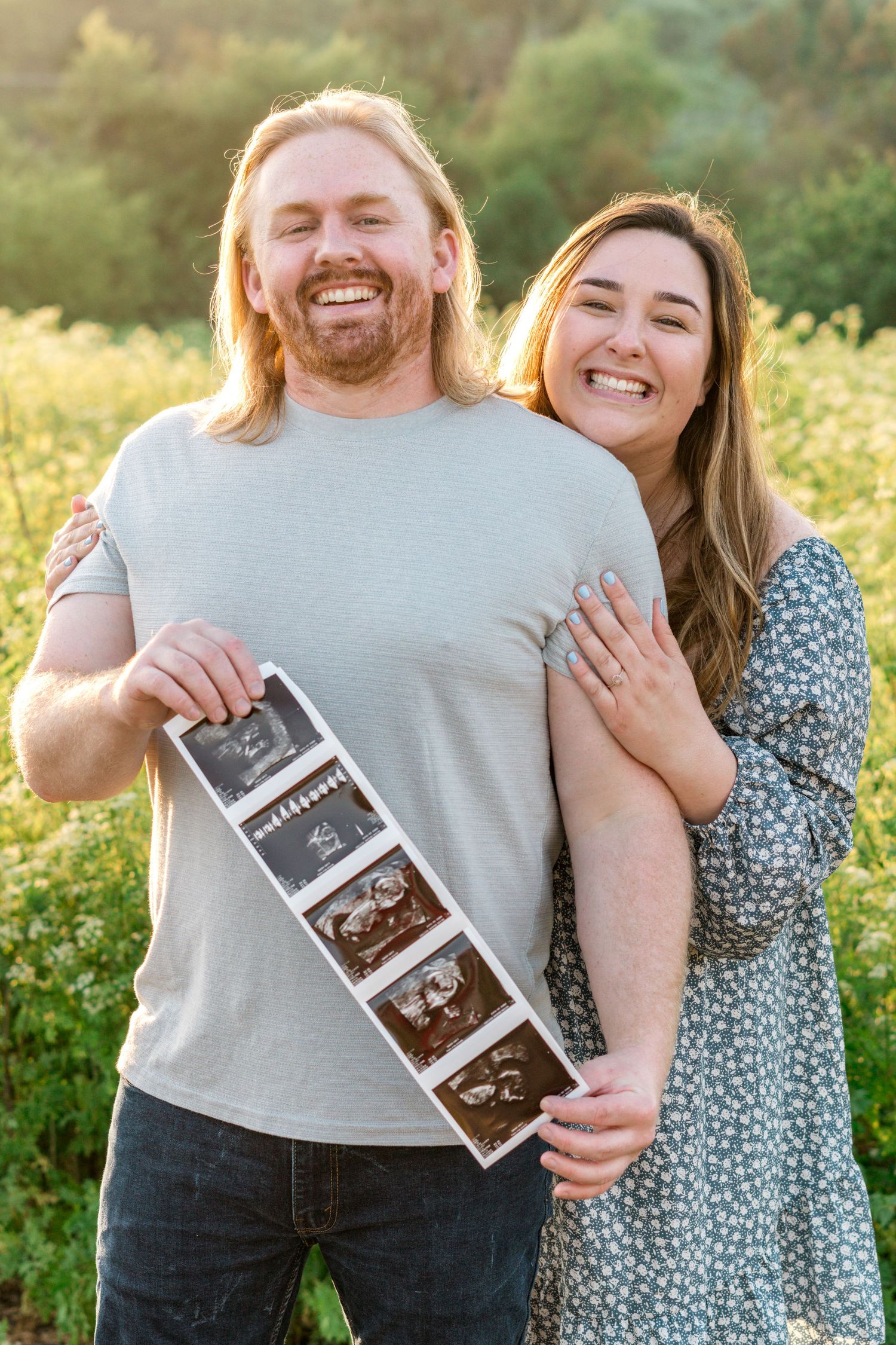 A couple stands in a sunlit field holding ultrasound photos while sharing joyful expressions and embracing.
