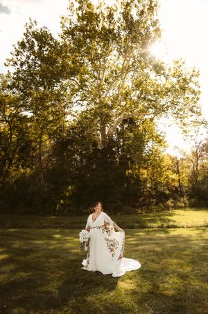 A bride in a white dress twirls in a sunlit meadow during golden hour with trees in the background.