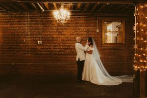 A romantic wedding couple dances in a rustic brick venue illuminated by warm string lights and chandeliers.