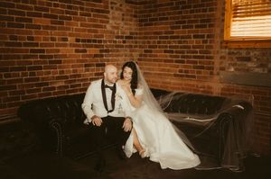 Newlywed couple sits intimately on brown leather couch against rustic brick wall backdrop in romantic setting.