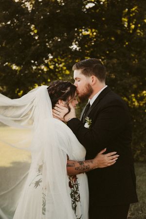 A romantic wedding couple embraces outdoors with the bride's veil flowing dramatically in the breeze.