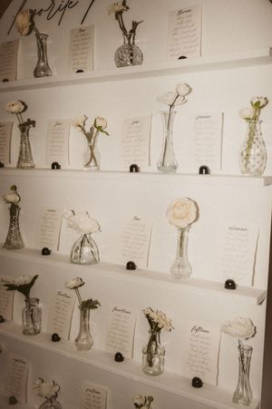Wedding seating chart display with small vases and handwritten cards on white shelves.