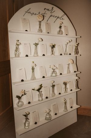 A wedding seating chart display featuring small glass vases with single flowers and name cards on white shelves.