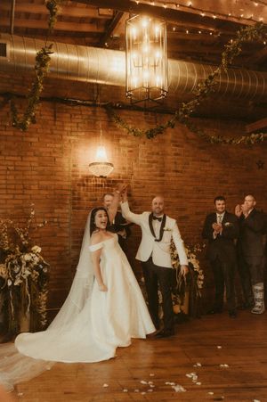 Wedding couple dances in a rustic brick-walled venue illuminated by string lights and chandeliers.