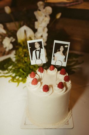 A white wedding cake decorated with raspberries and small photo toppers on a rustic wooden surface.