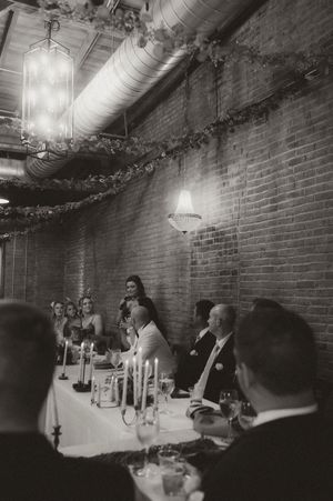 Elegant reception dinner in a rustic brick-walled venue with exposed ductwork and hanging chandeliers.