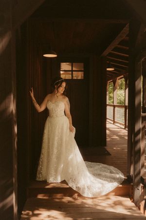 A white wedding dress with intricate lace details is illuminated by natural light streaming through windows in a rustic hallway.