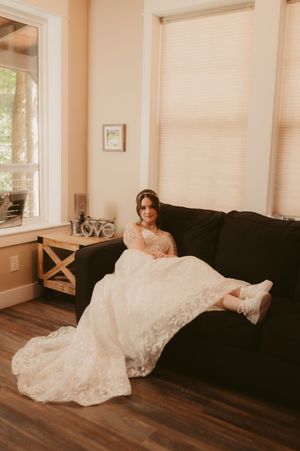 A bride in a white lace wedding dress lounges elegantly on a dark couch in a sunlit room.
