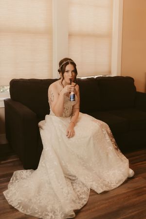 A bride in a white lace wedding dress sits on hardwood floors while holding a phone.
