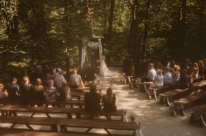 A rustic outdoor wedding ceremony in a sunlit forest with wooden benches and a white arch decorated with fabric.