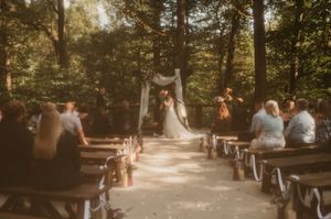 Wedding ceremony scene in a sunlit forest with guests seated on wooden benches.
