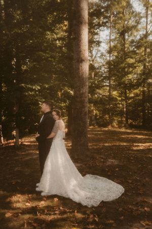 A bride in a flowing white gown poses dramatically in a sunlit forest setting.