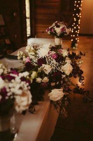 Elegant floral arrangements with white and purple blooms decorate a rustic wedding venue with twinkling lights.