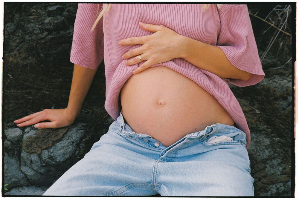 Pregnant woman in pink sweater and jeans sitting on rocks, cradling her baby bump outdoors.