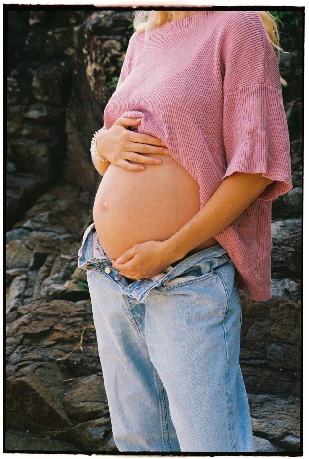 Pregnant woman in pink ribbed top and unbuttoned jeans cradling baby bump outdoors near stone wall.