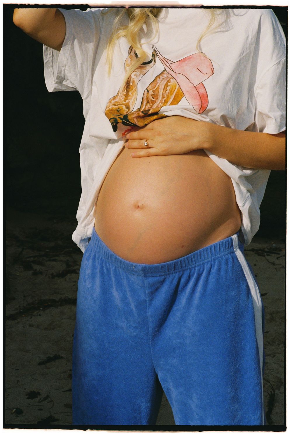 Pregnant woman lifting white graphic t-shirt to reveal baby bump, wearing blue velvet sweatpants outdoors.