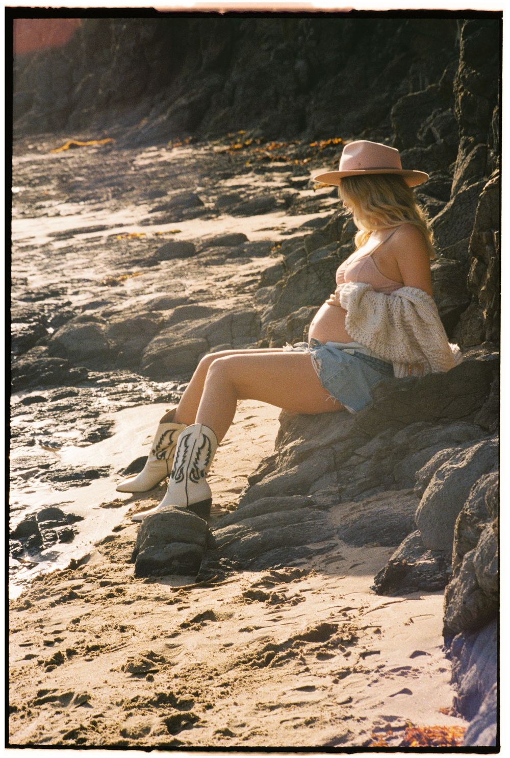 Pregnant woman in boho style sitting on beach rocks wearing cowboy boots, denim shorts, and a wide-brim hat.