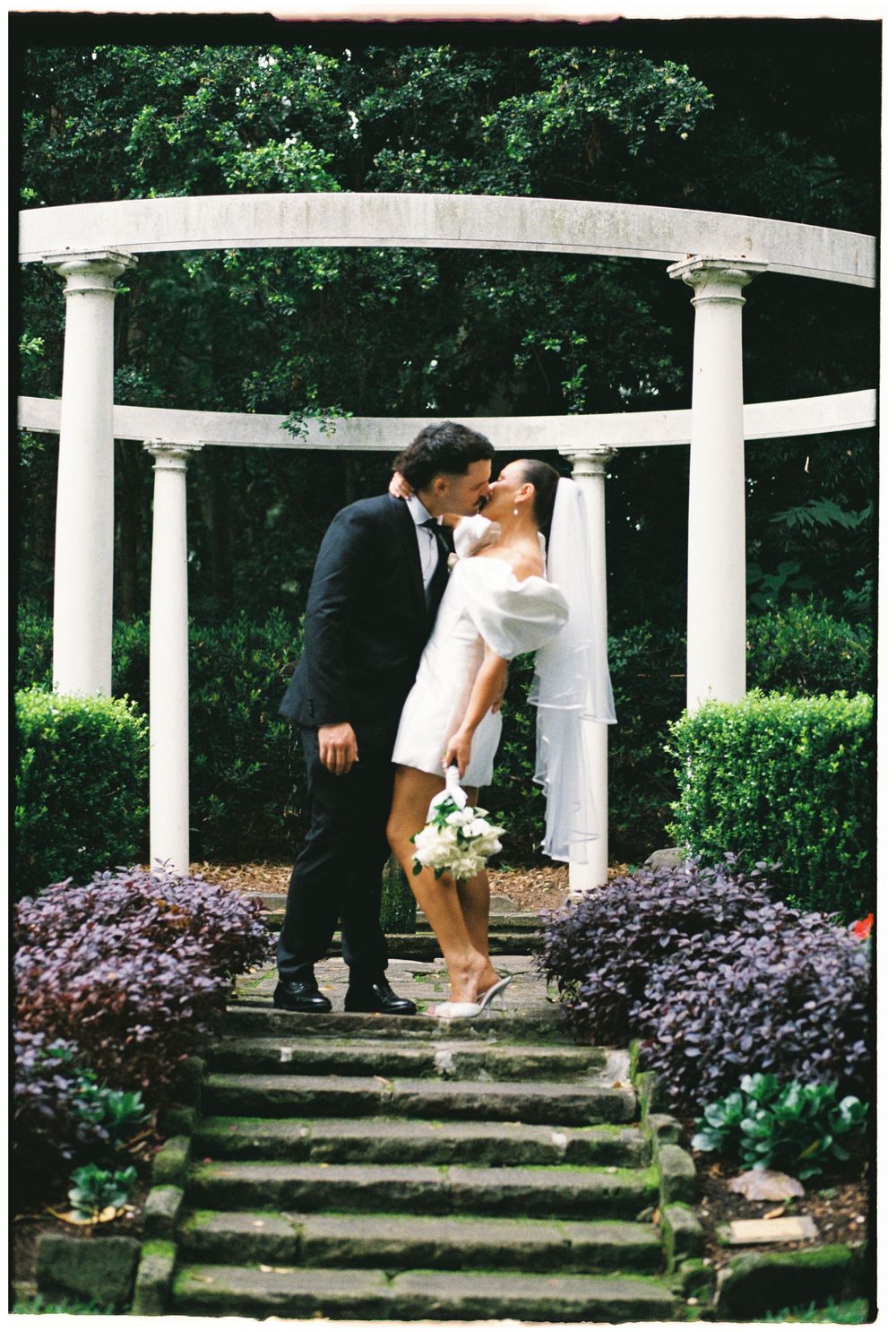 Bride and groom share a kiss on stone steps beneath a white columned garden gazebo surrounded by lush greenery.
