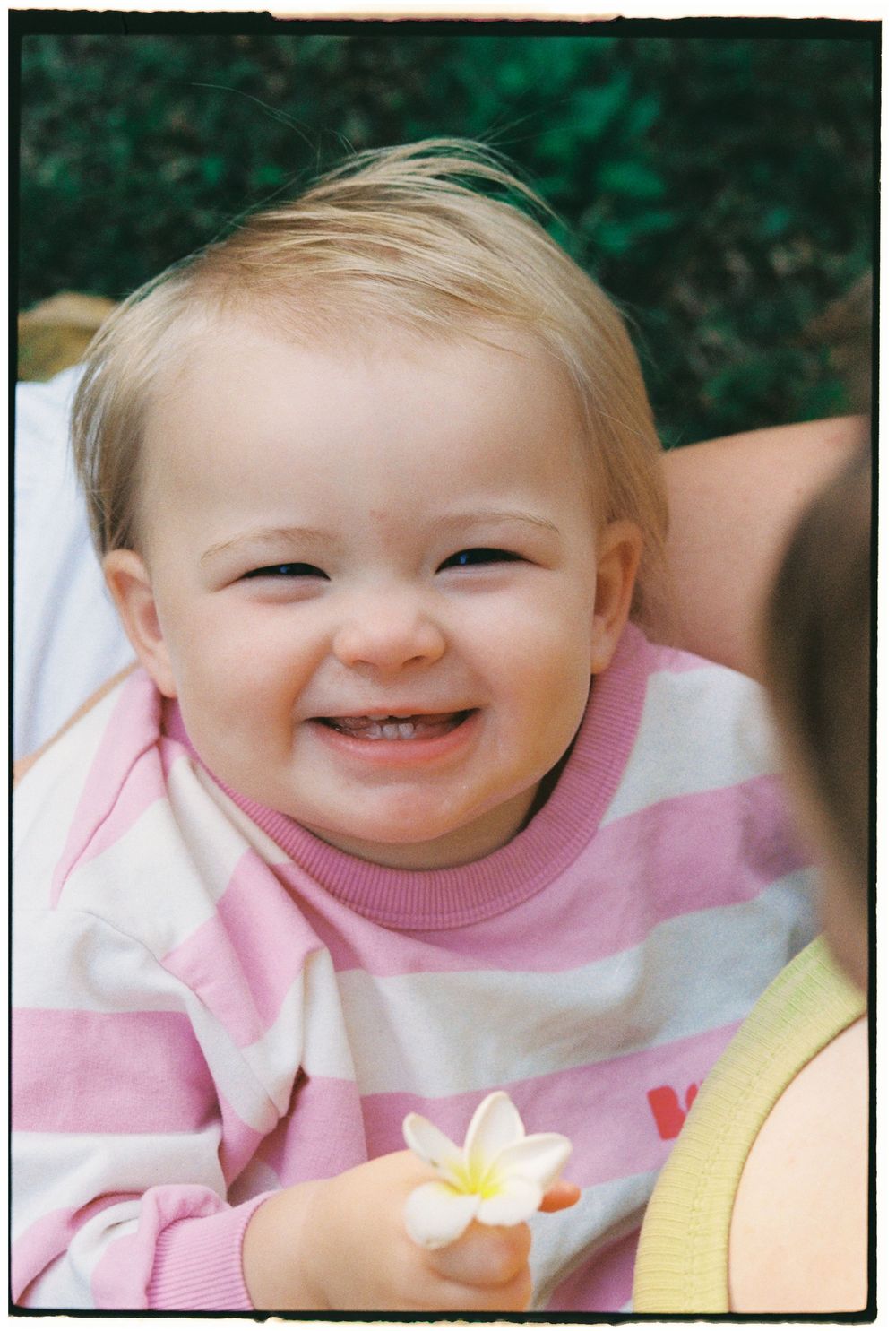 Smiling baby girl in pink striped shirt holding a white flower outdoors.