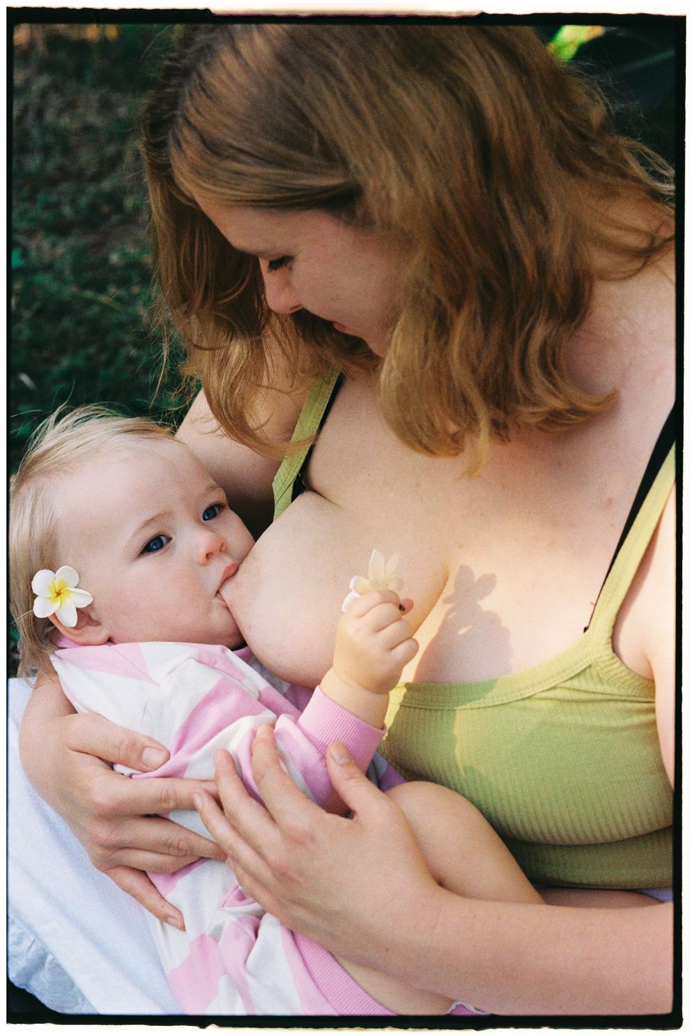 A mother breastfeeding her baby girl outdoors, with the infant wearing a white flower in her hair.