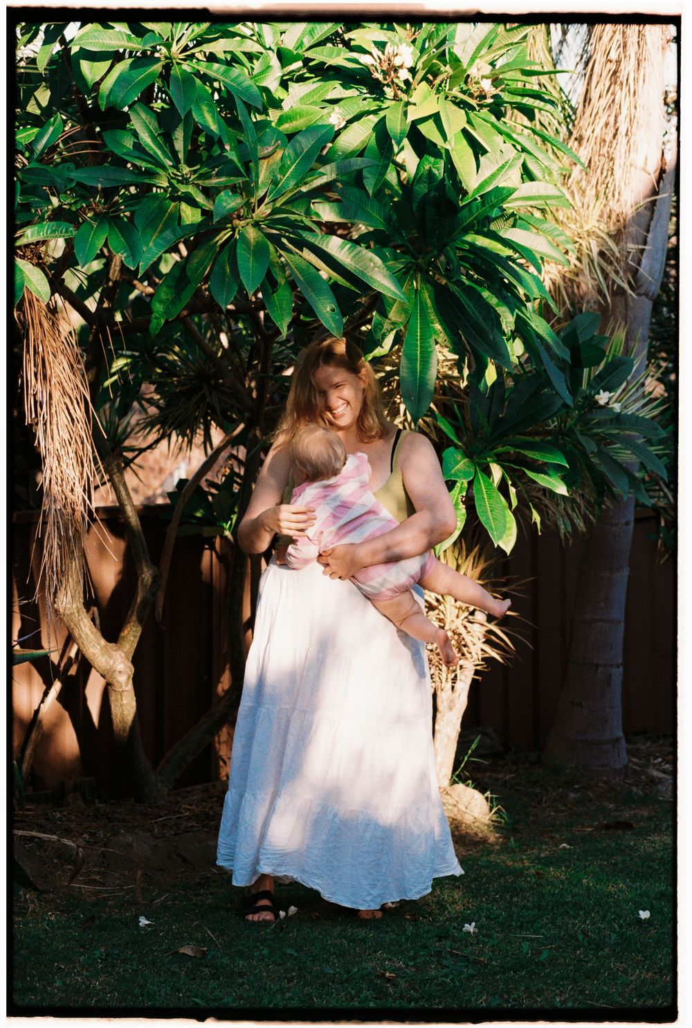 A mother tenderly holds her child outdoors near lush tropical plumeria trees in warm sunlight.