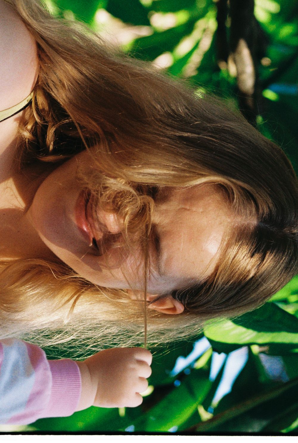 A young girl with long blonde hair holds a thin stick among lush green tropical leaves in sunlight.