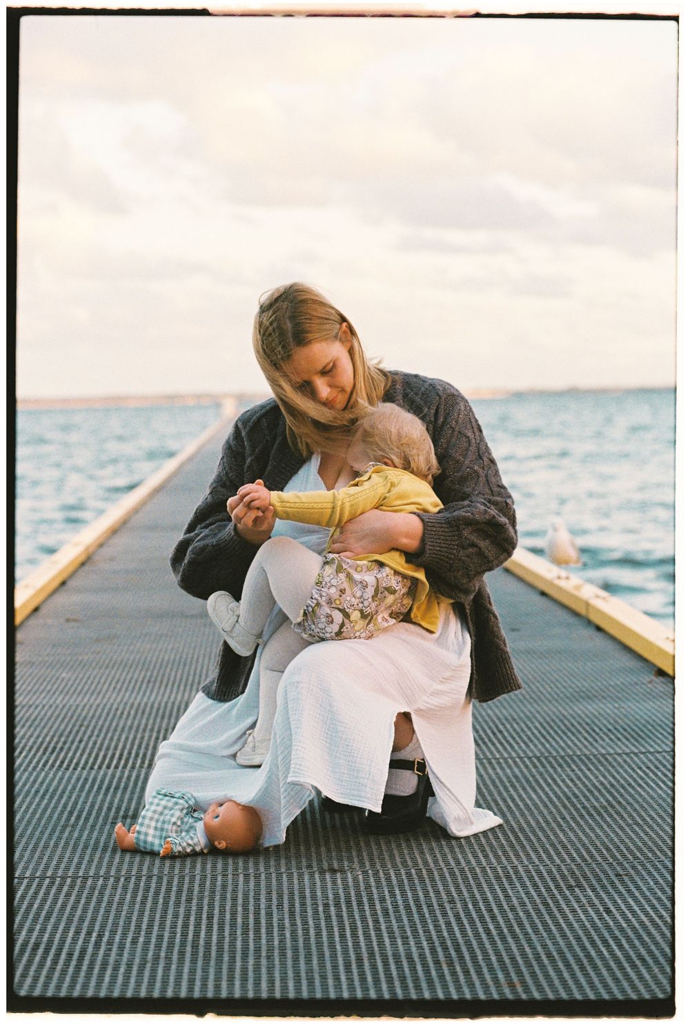 Mother kneeling on a seaside pier, tenderly holding her baby in a yellow sweater on a cloudy day.