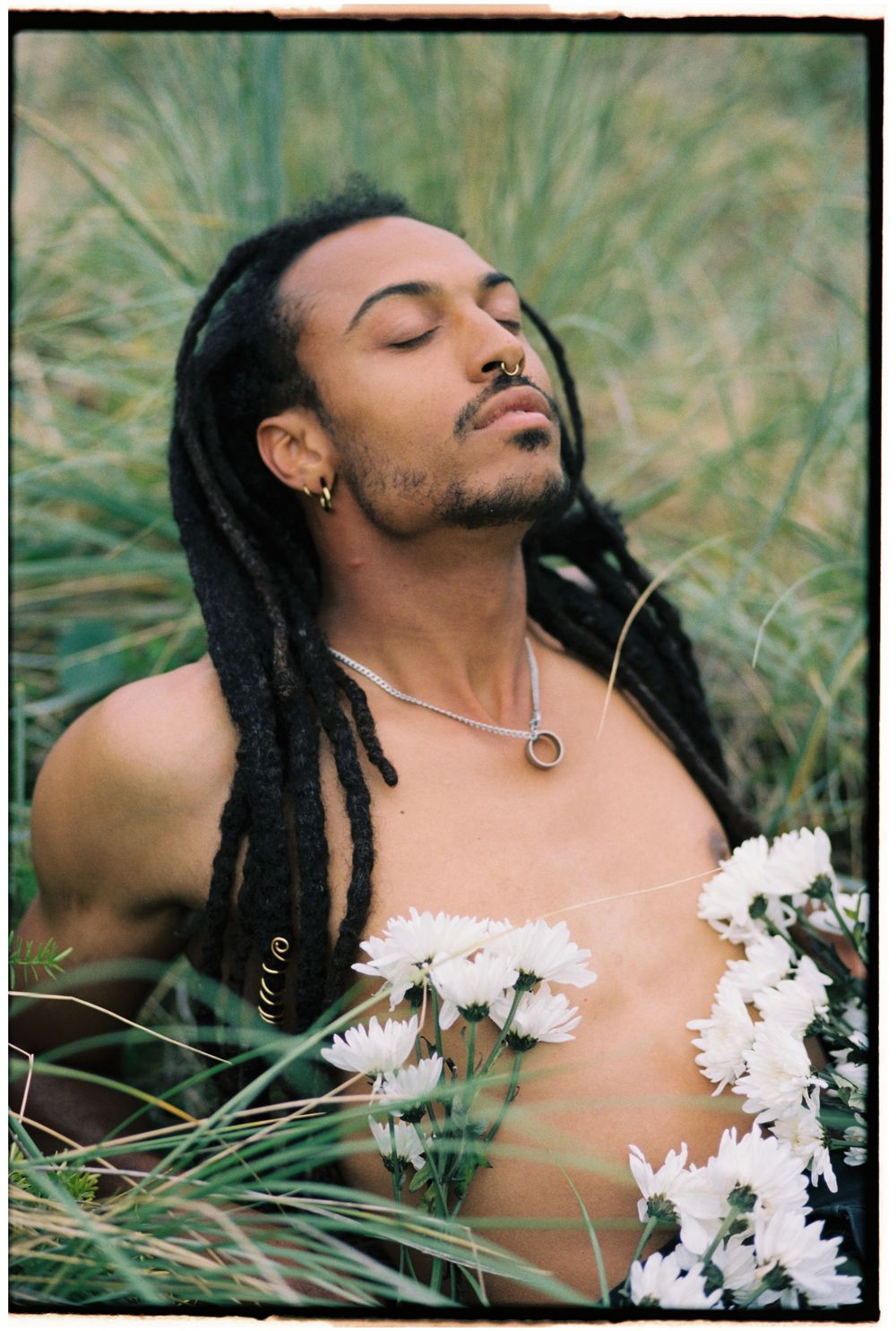 A person with dreadlocks lies peacefully in tall grass with white daisies scattered across their chest.