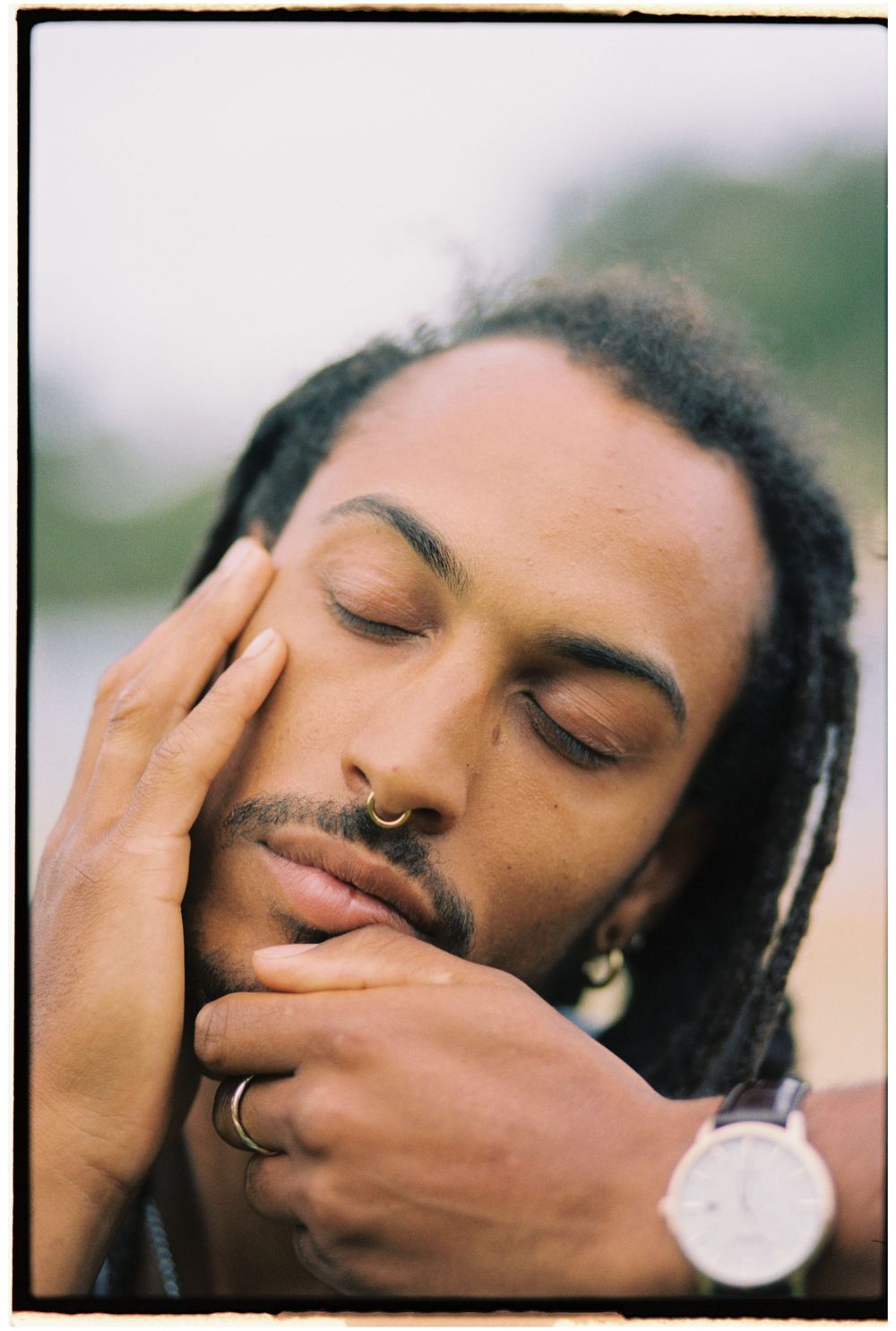 Close up portrait with eyes closed, wearing nose ring and rings, with dreadlocks against blurred background.